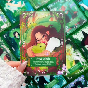 Peut inclure: Une carte de tarot avec une bordure verte, repr&eacute;sentant une femme avec des lunettes et une grenouille verte. La carte pr&eacute;sente des motifs de croissant de lune et de feuilles. Le texte sur la carte indique "frog witch" et "time to move to the next pond. let go of what no longer serves you. take a leap of faith."