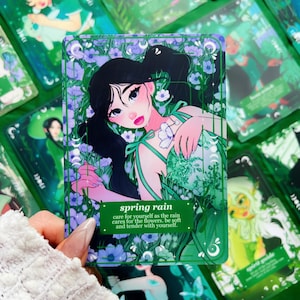 Peut inclure: Une carte de tarot avec une illustration florale d'une femme aux cheveux noirs. La carte a une bordure verte et le texte "spring rain" avec un message de soins personnels. D'autres cartes avec des designs similaires sont visibles.