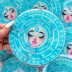 May include: A blue and white circular divination board with a stylized illustration of a woman's face. The board features the letters of the alphabet, numbers 1-9 and 0, and the words "yes", "no", "maybe", and "rephrase".