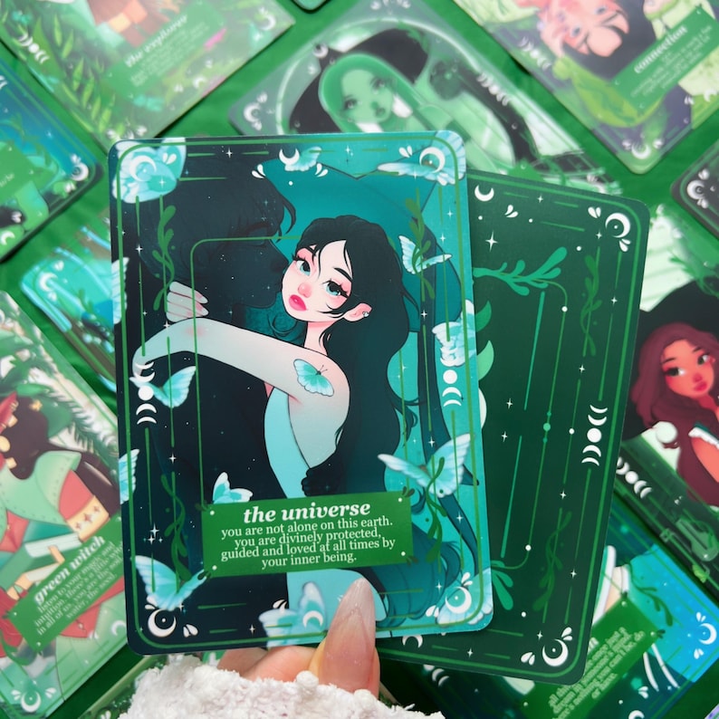 Peut inclure: Une carte de tarot verte et turquoise repr&eacute;sentant une femme aux longs cheveux noirs et des papillons. La carte indique "l'univers" avec un message inspirant. D'autres cartes sont visibles en arri&egrave;re-plan.