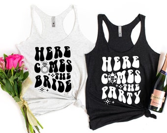 Puede incluir: Dos camisetas sin mangas blancas con texto negro. El texto de la camiseta de la izquierda dice "Here Comes the Bride" con un gráfico de anillo de diamantes. El texto de la camiseta de la derecha dice "Here Comes the Party" con un gráfico de bola de discoteca. Una botella de champán está en el fondo.