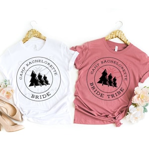 K&ouml;nnte beinhalten: Zwei T-Shirts mit einem schwarz-wei&szlig;en Grafikdesign. Das Design zeigt einen Kreis mit dem Text "Camp Bachelorette" und eine Silhouette von drei Kiefern. Ein Shirt ist wei&szlig; und das andere ist staubig rosa. Das wei&szlig;e Shirt sagt "Bride" und das staubig rosa Shirt sagt "Bride Tribe".