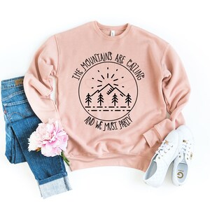May include: A blush pink sweatshirt with a black graphic print that reads "The mountains are calling and we must party". The graphic features a mountain range, a sun, and trees.