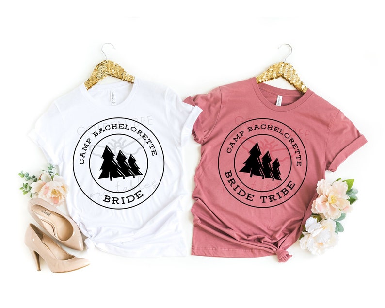 K&ouml;nnte beinhalten: Zwei T-Shirts mit einem schwarz-wei&szlig;en Grafikdesign. Das Design zeigt einen Kreis mit dem Text "Camp Bachelorette" und eine Silhouette von drei Kiefern. Ein Shirt ist wei&szlig; und das andere ist staubig rosa. Das wei&szlig;e Shirt sagt "Bride" und das staubig rosa Shirt sagt "Bride Tribe".