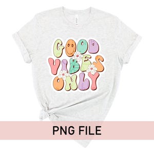 May include: A light grey t-shirt with a graphic print of the text "Good Vibes Only" in colorful, retro-style lettering with flowers and smiley faces.