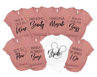 May include: A collection of eight t-shirts, primarily in a dusty rose color, with black text. One white t-shirt features a Mickey Mouse head design with the word "Bride." The other shirts have phrases like "Tale as Old as Wine" and "Bibbidi Bobbidi Booze."