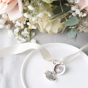 May include: A silver locket with a photo of a couple inside, open and resting on a white plate. The locket is attached to a white satin ribbon and is surrounded by a bouquet of white and pink roses.