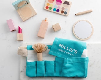 Pode incluir: Uma bolsa de maquiagem de tecido azul turquesa com uma etiqueta branca que diz "Millie's Make Up Kit". A bolsa tem vários bolsos e está cheia de itens de maquiagem de madeira para crianças, incluindo um pincel, batom, uma paleta de sombras para os olhos e um pequeno espelho.