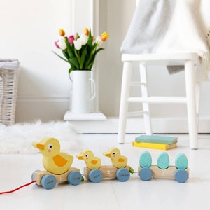 May include: A wooden toy train with three yellow duck carriages and a blue carriage with three green eggs. The train is on a white wooden floor. The carriages are numbered 1, 2, and 3. The duck carriages have the names Oscar, Maisie, and Tom on them. The blue carriage has the number 3 on it.