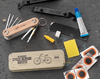 May include: A vintage-style metal tin with the text "WELDTITE CYCLE REPAIR OUTFIT for all Cycles ESTABLISHED 1940 BRITISH MADE" and a black and white illustration of a bicycle. The tin is open and contains a variety of tools, including a multi-tool with a wooden handle, a tire lever, a patch kit, and a tube of glue.