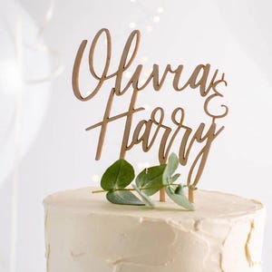 Può includere: Un topper per torta in legno con i nomi "Olivia & Harry" scritti in corsivo. Il topper è decorato con un rametto di verde.