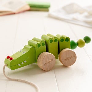 May include: A green wooden pull toy shaped like an alligator with a red nose and white teeth. The toy has wheels with the names "HARRY" and "19.09.2019" engraved on them.