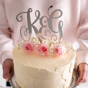 Peut inclure: Un gâteau blanc avec un glaçage au beurre crème décoré de roses roses et de gypsophile. Le gâteau est surmonté d'un topper de gâteau en acrylique miroir argenté avec les lettres "K & G" en écriture cursive.