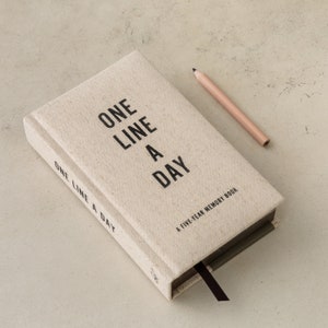 Op de afbeelding: Een beige, met linnen bedekt tijdschrift met de tekst "ONE LINE A DAY" in zwart. Een lichtroze potlood ligt naast het boek. Het tijdschrift heeft ook een zwart lint en de tekst "A FIVE-YEAR MEMORY BOOK".