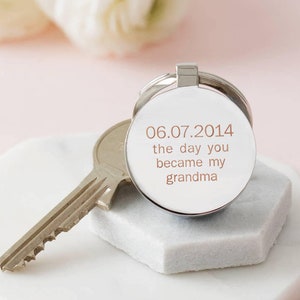 May include: Silver keychain with a round charm engraved with the date "06.07.2014" and the text "the day you became my grandma".