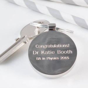May include: Silver keychain with a round engraved tag that reads "Congratulations! Dr Katie Booth BA in Physics 2015".