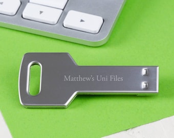 May include: A silver key-shaped USB drive with the text "Matthew's Uni Files" engraved on it. The drive is lying on a green surface next to a silver keyboard.