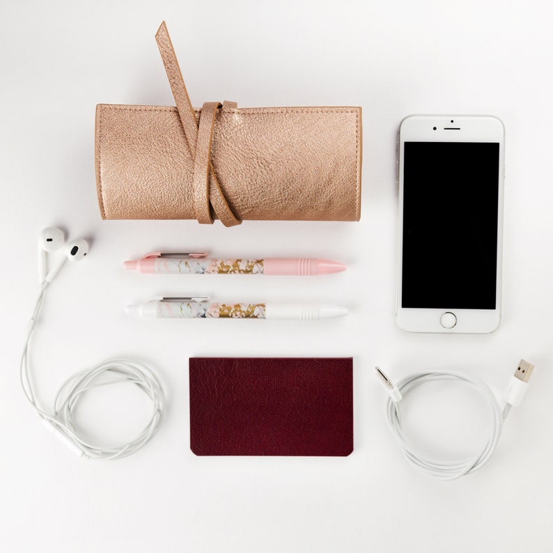 May include: A flat lay of a gold leather pencil case, two pens with floral designs, a burgundy leather notebook, a white phone with a black screen, and white earbuds.