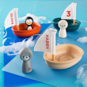 May include: Three toy boats with names Olivia, Harry, and the number 3 on the sails. The boats are made of wood and plastic and are floating in a blue tub of water. There is a penguin and a seal figurine in the boats.