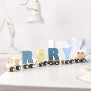 May include: A wooden toy train spelling out the name "RORY". The train cars are in shades of blue and green. The wooden train has wheels and the letters are attached to the carriages. The toy is made of wood and is on a white surface.