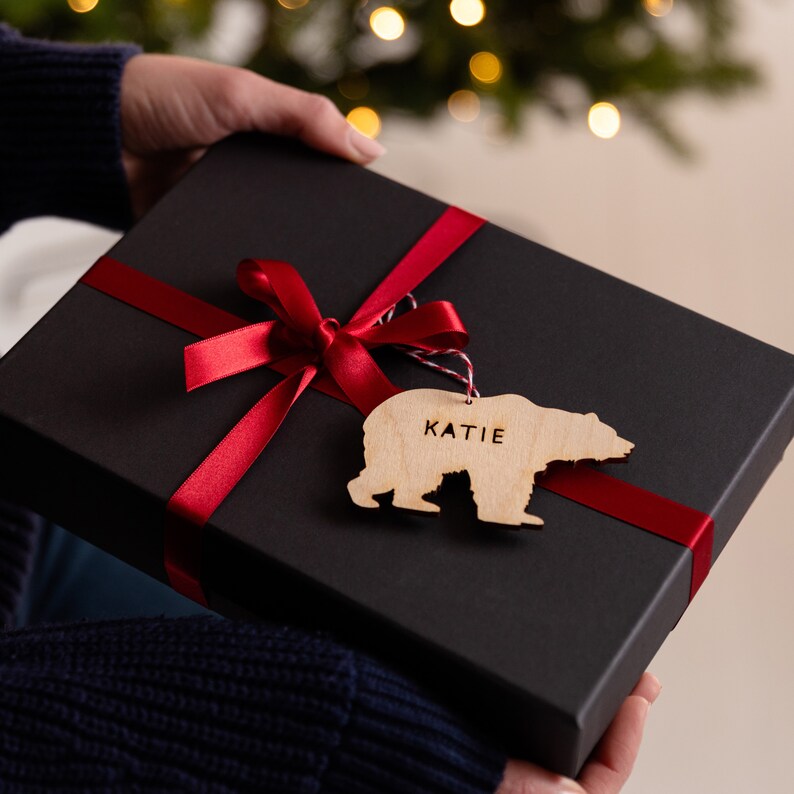 May include: A black gift box tied with a red ribbon and bow. A wooden bear-shaped ornament with the name "KATIE" is attached. The background features a blurred Christmas tree with bokeh lights.
