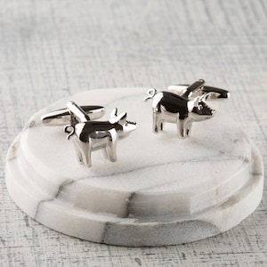 May include: A pair of silver pig cufflinks. The cufflinks are in the shape of a pig, with the pig's tail curled up. The cufflinks are on a white marble surface.