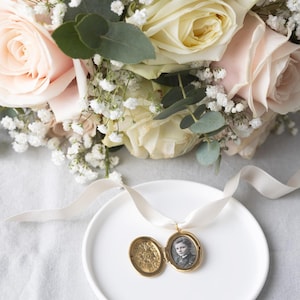 Può includere: Un medaglione d'oro con un piccolo ritratto all'interno, appoggiato su un piatto bianco con un nastro color crema. Un bouquet di rose rosa chiaro e crema e velo da sposa è sullo sfondo.