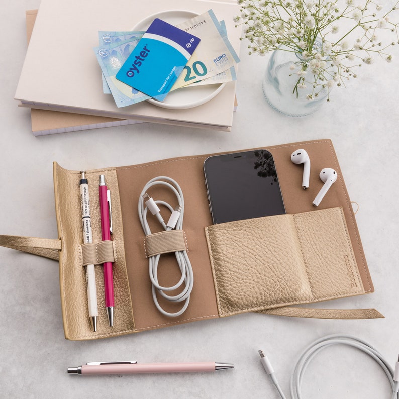 May include: A gold leather travel organizer with a phone pocket, two pen slots, a cable holder, and two white wireless earbuds. The organizer is open and laid flat on a white surface.