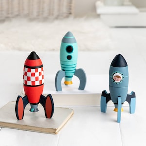 May include: Three wooden toy rockets in red, teal, and blue. The red rocket has a checkered pattern, the teal rocket has stripes, and the blue rocket features an astronaut in a window. Each rocket has grey fins and yellow accents, ideal for imaginative play.