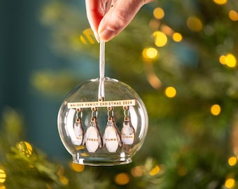 May include: A clear glass Christmas ornament with a wooden plaque that reads "Walker Family Christmas 2024". Inside the ornament are five wooden penguin ornaments with the names "Oscar", "Mummy", "Daddy", and two others.