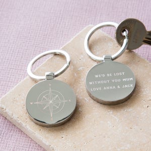 Könnte beinhalten: Silberner Schlüsselanhänger mit einem Kompass-Design auf der einen Seite und dem Text "We'd be lost without you Mum Love Anna & Jack" auf der anderen Seite.