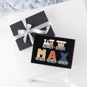 Op de afbeelding: Een zwarte cadeaudoos met een wit lint en strik. De doos is open en bevat een houten trein met de naam "MAX" gespeld in kleurrijke letters.