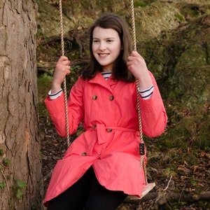 May include: A young girl wearing a pink raincoat with a belt sits on a rope swing in a wooded area.