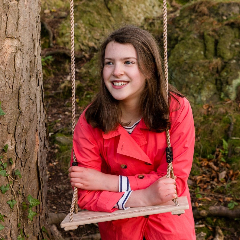 May include: A young person with long brown hair is sitting on a wooden swing with rope. They are wearing a red jacket with a white stripe on the sleeve. The swing is hanging from a tree branch.