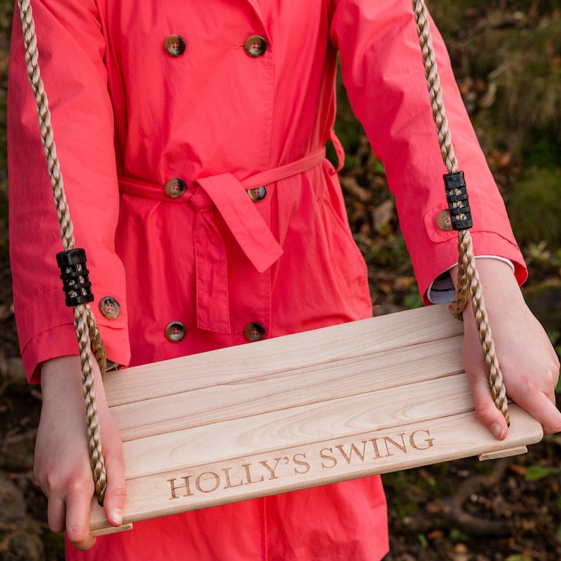 May include: A wooden swing set with the words "HOLLY'S SWING" engraved on the seat. The swing is made of natural wood and has a rope with a knot at the end for hanging.