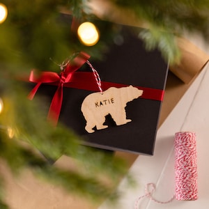 May include: A wooden bear-shaped ornament with the name "KATIE" is attached to a wrapped gift. The gift is a black box with a red ribbon. The ornament hangs from a red and white string. The background includes a Christmas tree and wrapping paper.