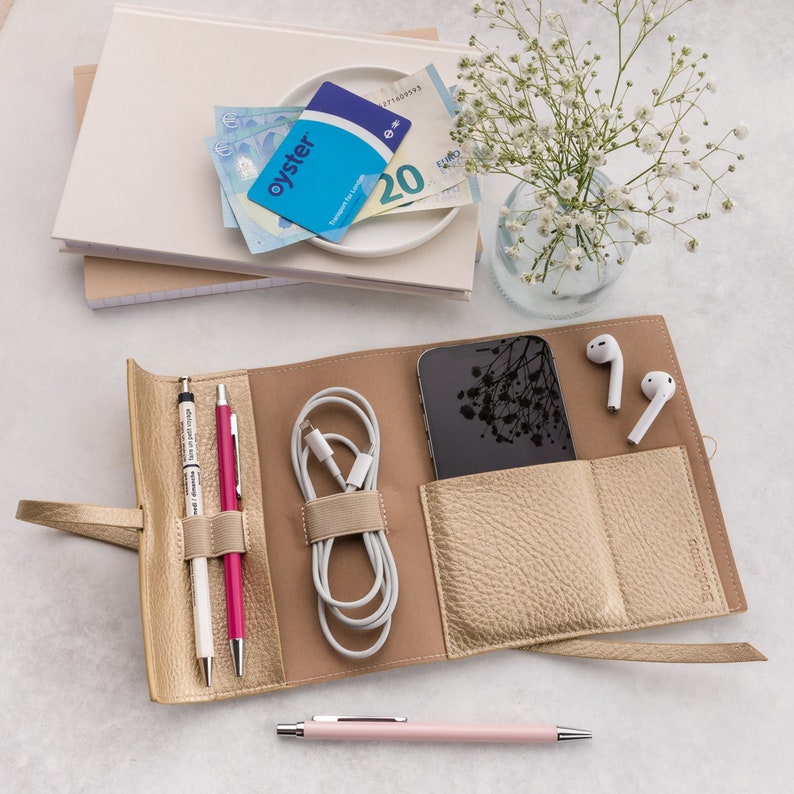 May include: A gold-colored leather travel organizer with multiple pockets and compartments. It includes a phone pocket, two pen holders, a pocket for a charging cable, and two pockets for earbuds. The organizer is open and laid flat on a white surface.