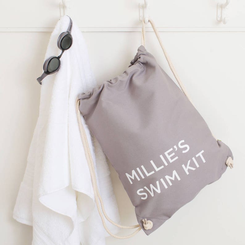 Op de afbeelding: Een grijze trekkoordtas met witte tekst die "Millie's Swim Kit" zegt. De tas hangt aan een witte haak met een witte handdoek en een paar zwarte zwembrillen.