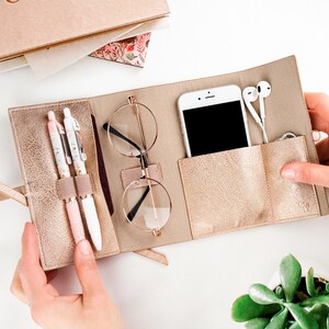 May include: A gold-colored leather travel case with a zipper closure. The case has multiple compartments for storing items such as a phone, glasses, pens, and earbuds. The case is open and the items are visible.