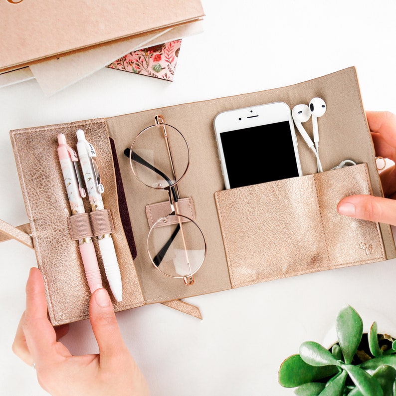May include: A gold-colored leather travel case with a zipper closure. The case has multiple compartments for storing items such as a phone, glasses, pens, and earbuds. The case is open and the items are visible.