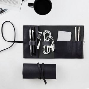 May include: Black leather roll-up case with multiple compartments for carrying pens, pencils, a compass, and earbuds. The case is rolled up and tied with a black leather strap.
