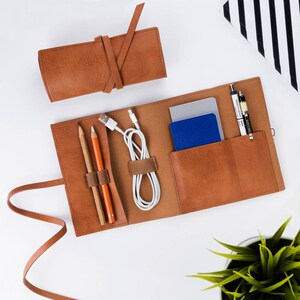 May include: A brown leather travel organizer with multiple pockets and compartments. It includes a zippered pouch, two pencil holders, and a space for a phone or other small electronics. The organizer is open and laid flat on a white surface.
