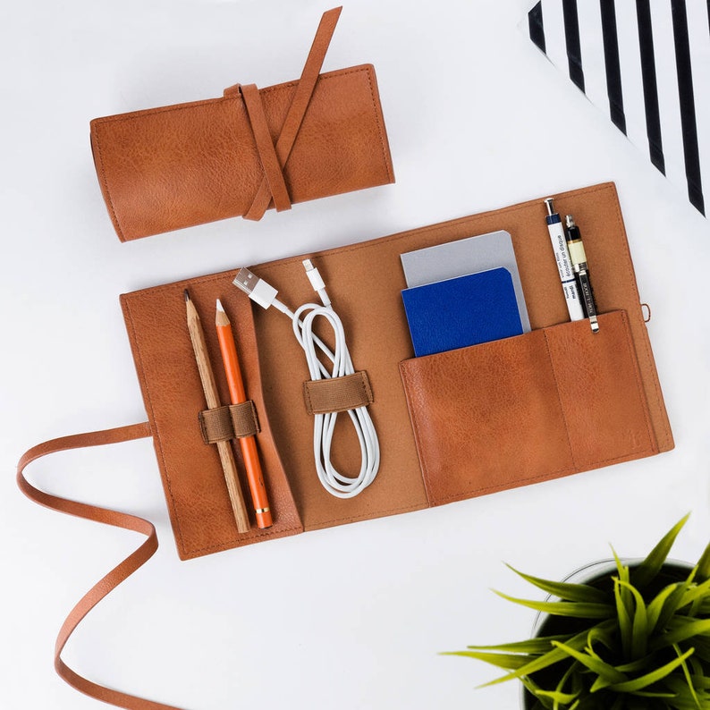 May include: A brown leather travel organizer with multiple pockets and compartments. It includes a zippered pouch, two pencil holders, and a space for a phone or other small electronics. The organizer is open and laid flat on a white surface.