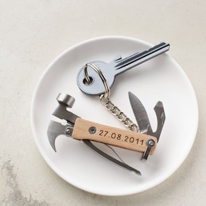 Può includere: Una chiave d'argento con un portachiavi attaccato a un multiutensile con un manico in legno. Il manico è inciso con la data "27.08.2011".