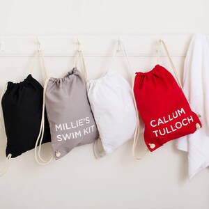 Op de afbeelding: Vier trekkoordtassen met gepersonaliseerde namen. De tassen zijn zwart, grijs, wit en rood. De tekst op de tassen luidt "Millie's Swim Kit" en "Callum Tulloch".