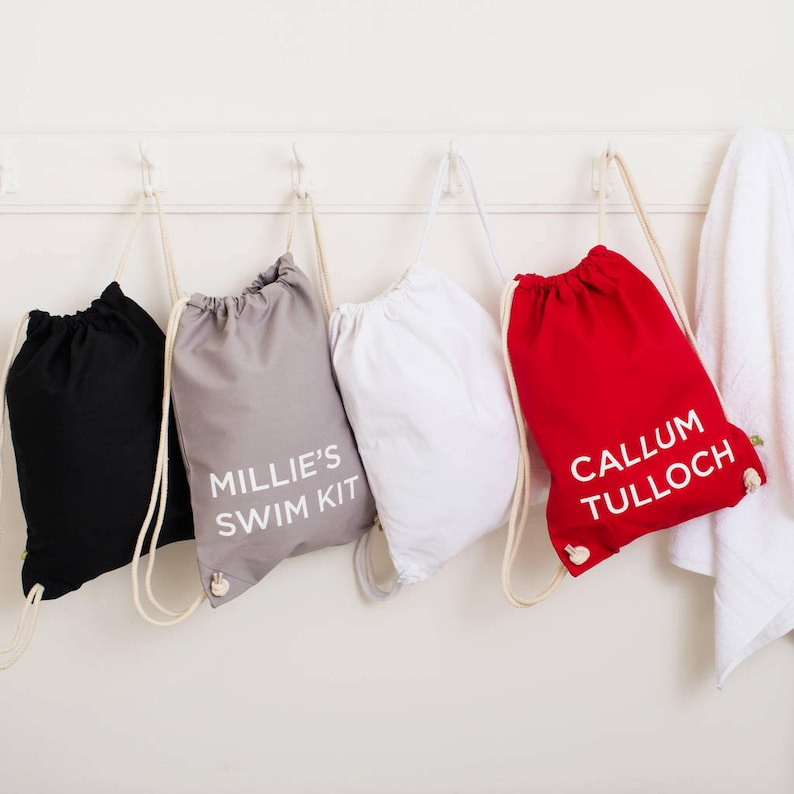 Op de afbeelding: Vier trekkoordtassen met gepersonaliseerde namen. De tassen zijn zwart, grijs, wit en rood. De tekst op de tassen luidt "Millie's Swim Kit" en "Callum Tulloch".
