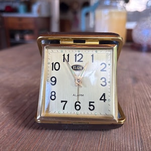 Puede incluir: Un reloj despertador de viaje cuadrado Elgin con marco de metal dorado y estuche abatible de color marrón oscuro. La esfera tiene un fondo dorado cepillado, números arábigos negros del 1 al 12, manecillas doradas y la palabra ALARM impresa.