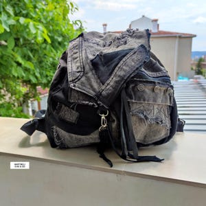 May include: A distressed black and gray denim backpack with multiple pockets and straps. The backpack features a flap closure, metal hardware, and visible stitching details. The bag has a rugged, worn aesthetic.