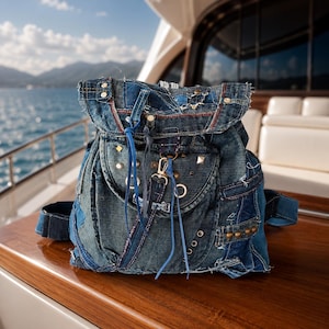 Op de afbeelding: Een patchwork rugzak van denim in verschillende tinten blauw, voorzien van gerafelde randen, meerdere metalen studs, zilverkleurige ringen, een voorflap met trekkoordsluiting en een kleine metalen karabijnhaak bevestigd aan een ring.
