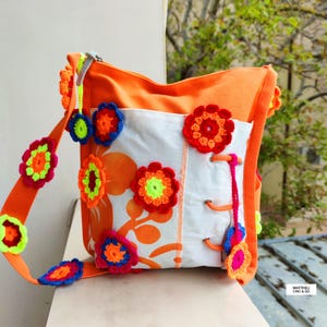 K&ouml;nnte beinhalten: Eine orangefarbene Umh&auml;ngetasche mit einer wei&szlig;en Frontplatte mit orangefarbenen Blumenmustern. Die Tasche ist mit bunten, geh&auml;kelten Blumenapplikationen in Rot, Blau und Gelb verziert. Der Riemen hat auch geh&auml;kelte Blumen.